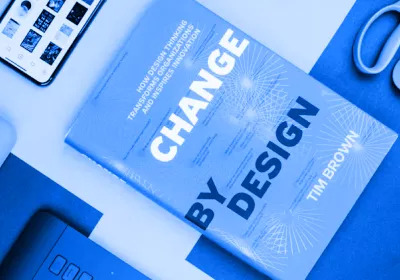 A top-down, blue-toned flat lay featuring the book "Change by Design" by Tim Brown. Surrounding the book are creative workspace essentials including a smartphone showing a grid of images, a pair of scissors, and a designer’s tablet.
