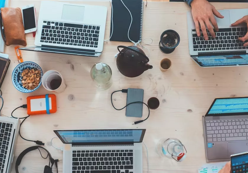 An overhead view of a busy, light-colored wooden office desk where several people are working on multiple laptops. The workspace is filled with creative tools and tech accessories, including tangled charging cables, a pair of headphones, an external hard drive, a bowl of nuts, a glass water bottle, and a teapot.