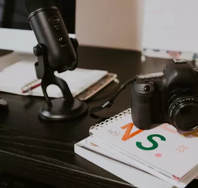 A high-angle shot focusing on a desk surface set up for media production. A professional studio microphone stands upright next to a DSLR camera with the lens cap attached, resting on top of a stack of notebooks and a binder labeled "NS 2020."