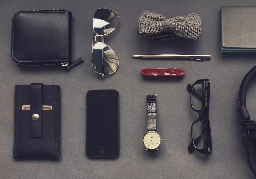 A neatly organized flat lay of men's style accessories against a dark grey background. The items include a black leather wallet, a pair of aviator sunglasses, a tweed bowtie, a sleek metal pen, a smartphone, a wristwatch, black-rimmed glasses, and a portion of a set of headphones.