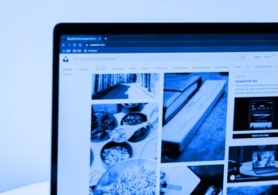 A close-up, blue-tinted shot of a laptop screen displaying the Unsplash website. The screen shows a grid of various high-quality photographic images, highlighting digital curation and product exploration.